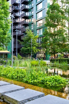 Stepping stones through an urban garden in Europe, with water, landscaped greenery and modern residential buildings surrounding the path