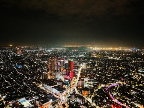 Aerial view of the illuminated cityscape of Surabaya at night, featuring glowing skyscrapers, dense urban infrastructure, and vibrant city lights in Surabaya, East Java, Indonesia.