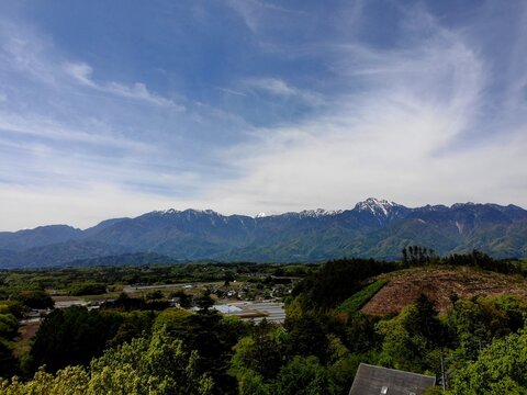 Aerial view of the Yatsugatake Mountains rising above lush green foothills and rural fields under a blue sky with wispy clouds in Chino, Nagano, Japan.