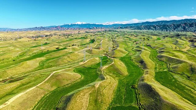 High angle view showcases rolling green hills stretching toward snow capped mountains under a clear blue sky, emphasizing vast natural beauty.