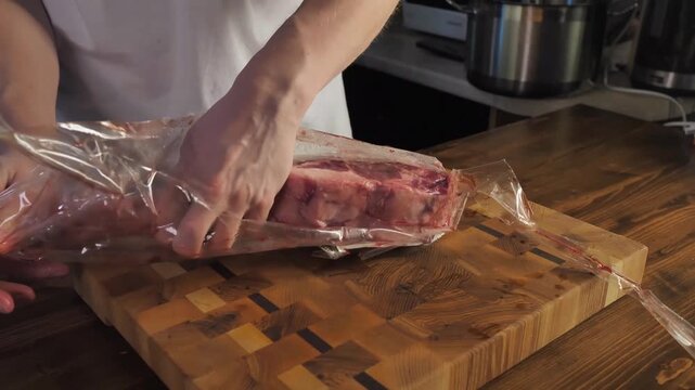 Hands lifting long strip of raw meat from clear plastic wrap on patterned wooden cutting board in warm home kitchen with stainless cookware softly blurred behind