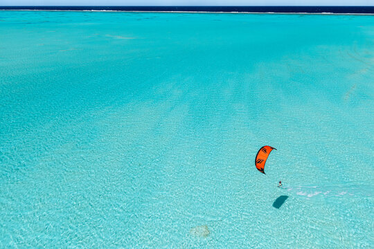 Aerial view of a kitesurfer with an orange kite gliding over the clear turquoise waters of the Aitutaki lagoon, Aitutaki, Cook Islands.