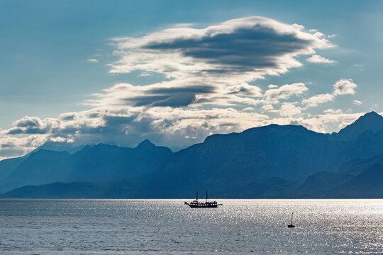 View of a small boat sailing on the calm sea with the majestic Taurus Mountains in the background under a dramatic cloudy sky in Antalya, Antalya, Turkey.