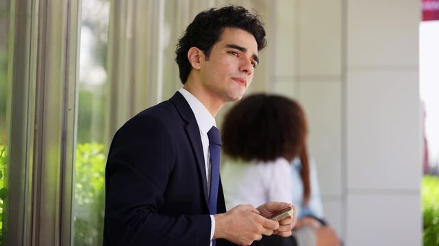 Businessman on a phone call at a covered waiting area, standing up and leaning in as he talks through a problem, a colleague texting behind him in soft focus on a busy workday
