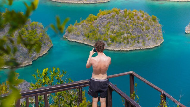 View of a man standing on a wooden viewing platform at Piaynemo taking a photo of the karst islands surrounded by turquoise water in Raja Ampat, Papua Barat, Indonesia.