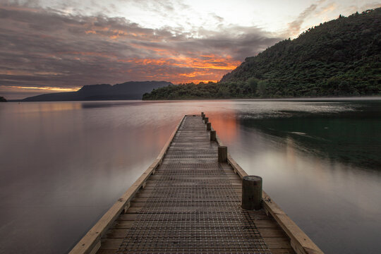 View of Lake Tarawera with a wooden jetty extending into the calm water at sunset, featuring soft clouds and distant hills in Rotorua, Bay of Plenty Region, New Zealand.