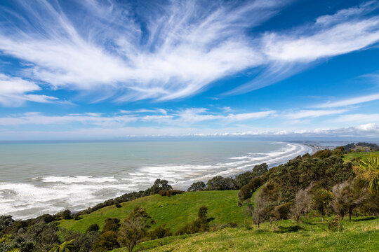 View of Ohope Beach with rolling green hills, crashing waves, and wispy clouds in the sky in Ohope, Bay of Plenty Region, New Zealand.