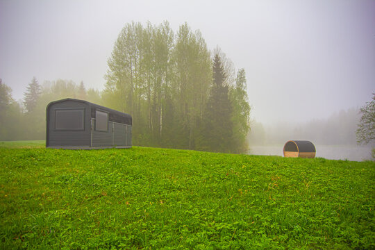 View of a modern rectangular cabin and a barrel sauna on a lush green meadow by a misty lake surrounded by trees in Latvia.