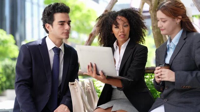 Laptop shared outdoors on a bench as a businessman and his teammates review a project together, coworking well beyond the office walls on a bright green urban campus