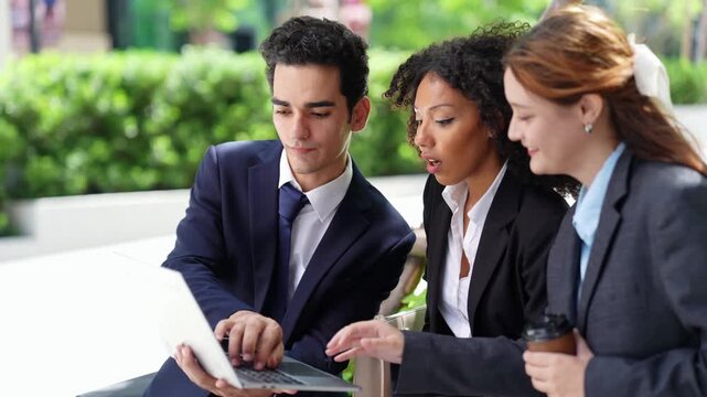Three colleagues cluster around one laptop screen outdoors, smiling as a result comes in, a candid teamwork moment on a landscaped corporate plaza in the morning sun