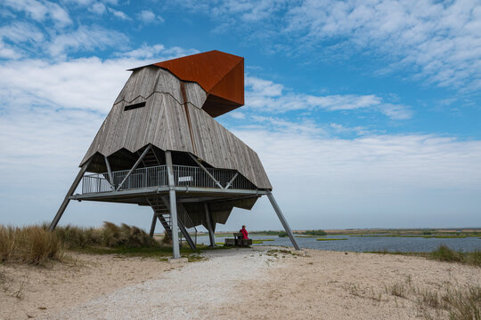 View of the Marker Wadden bird-watching tower with a person sitting on a bench nearby, set against a blue sky and water in Lelystad, Flevoland, Netherlands.
