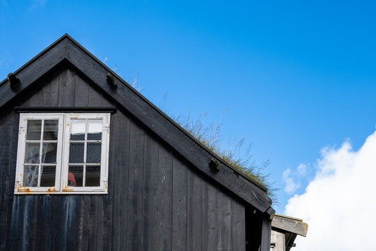 View of a traditional black wooden house with a white-framed window and a lush green turf roof under a clear blue sky in the Faroe Islands.