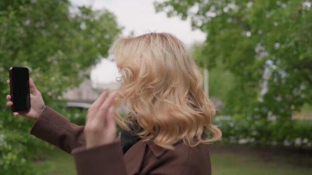 Blonde woman wearing brown coat holds up her smartphone to take photo of herself outdoors. Woman adjusts her hair while posing for selfie set against backdrop of green trees and distant