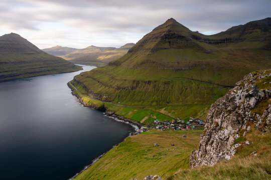 View of a small village nestled along the coast of a deep fjord, surrounded by steep, grass-covered mountains under a soft, cloudy sky in the Faroe Islands.