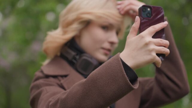 Lady holds up smartphone to take self portrait selfies outdoors. Woman adjusts hair and positions hand on device with greenery blurred in background selfie