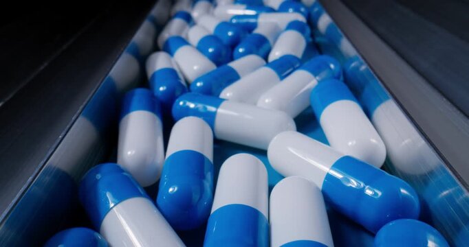 Tracking shot of blue and white capsules tumbling along stainless conveyor on pharmaceutical production line.