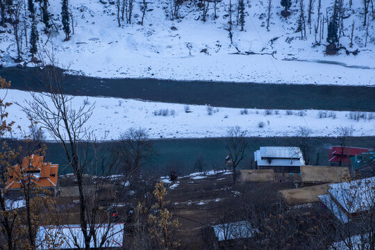 View of the village nestled in snow-covered mountains with a river flowing through the valley, featuring traditional houses and bare trees in Arang Kel, Kashmir, Pakistan.
