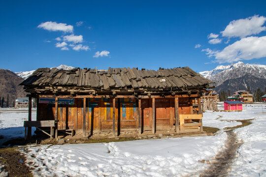 View of a traditional wooden house with a weathered roof set against a backdrop of snow-covered mountains in Arang Kel, Kashmir, Pakistan.