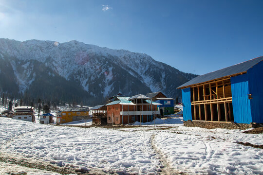 View of Arang Kel village with wooden houses and structures set against snow-covered mountains under a clear blue sky in Arang Kel, Kashmir, Pakistan.