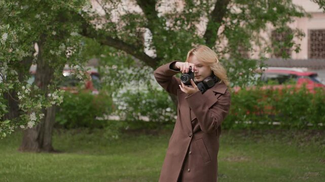 Lady stands outdoors holding digital camera framed from waist up. Woman wears long brown coat and has blonde hair. background features green trees with white flowers grassy ground distant