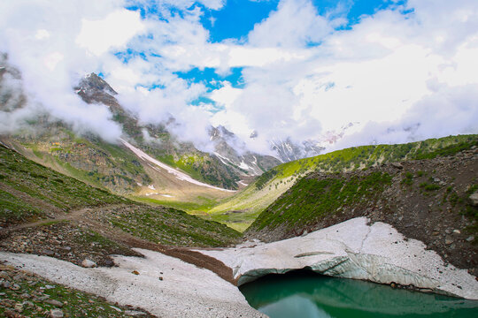 View of Rama Meadows featuring a glacial lake, snow-capped mountains, and lush green slopes under a cloudy sky in Astore, Gilgit, Pakistan.