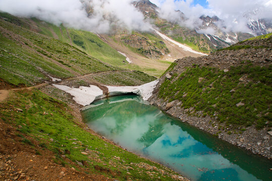 View of the turquoise waters of Rama Lake surrounded by snow-capped mountains, green meadows, and rocky slopes in Astore, Gilgit, Pakistan.