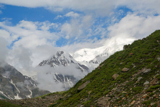 View of the snow-capped peaks of Rama Meadows rising above lush green slopes and rocky terrain under a cloudy blue sky in Astore, Gilgit, Pakistan.