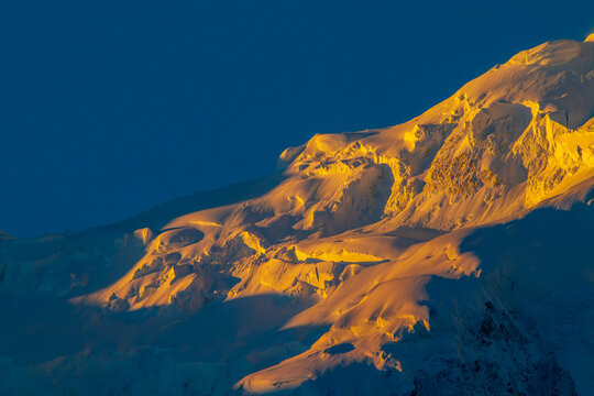 View of the snow-covered peaks of Nanga Parbat illuminated by golden sunlight against a deep blue sky in Fairy Meadow, Gilgit, Pakistan.