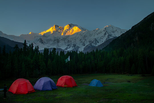 View of Nanga Parbat peak illuminated by golden sunrise light behind a campsite with colorful tents and a pine forest in Fairy Meadow, Gilgit, Pakistan.