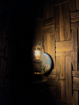 Dimly lit traditional oil lamp with a glass chimney casting a faint warm glow against a dark woven wooden wall, creating a moody and nostalgic atmosphere in quiet low light.
