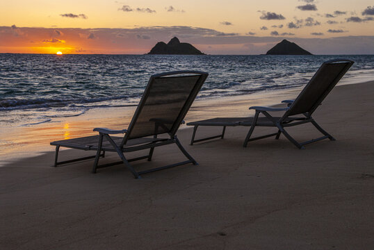 View of two lounge chairs on the sand at Lanikai Beach during sunset with the Mokulua Islands in the background in Kailua, Hawaii, United States.