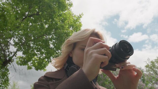 Woman holds digital camera in outdoor park setting. Woman adjusts settings on her handheld device while wearing headphones around her neck suggesting woman is preparing to take photos
