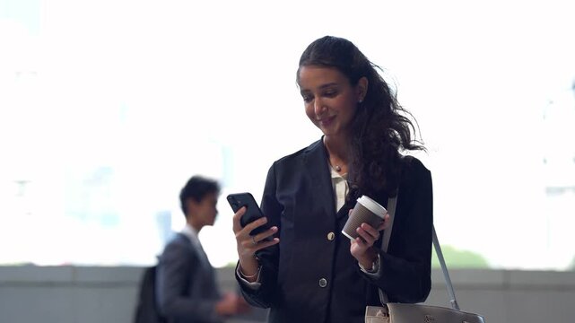 Latina businesswoman talks on her mobile phone outdoors, then smiles as she scrolls messages and sips takeaway coffee while a male colleague walks past out of focus in the background