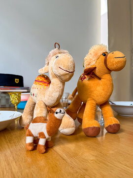 Family of plush camel toys in cream and orange tones, with two large adults and a small baby camel arranged together on a wooden table, symbolizing togetherness and childhood play.