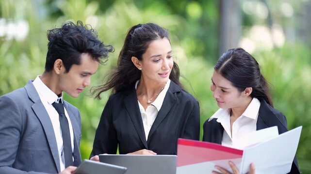 Tight three-shot of young business colleagues reviewing a red report folder alongside a tablet and a laptop outdoors, heads close together as they check the figures in a green garden