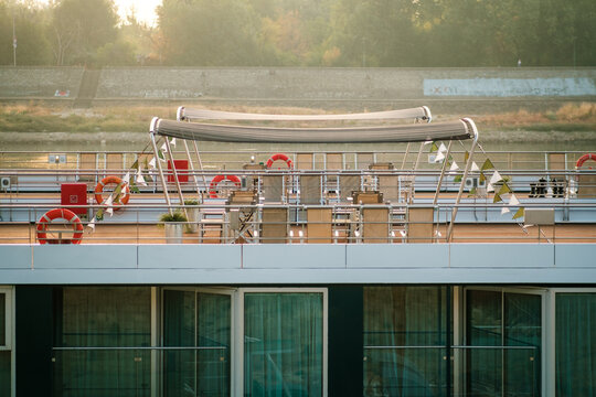 View of the sunlit deck of a river cruise ship with tables, chairs, and lifebuoys, docked along the river in Novi Sad, Vojvodina, Serbia.