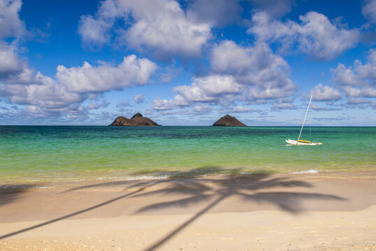 View of Lanikai Beach with turquoise water, a small sailboat, and the Mokolua Islands in the distance under a blue sky with clouds in Kailua, Hawaii, United States.