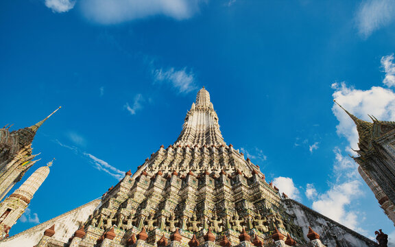 View of the ornate central prang of Wat Arun temple decorated with colorful porcelain mosaics under a bright blue sky in Wat Arun, Bangkok, Thailand.