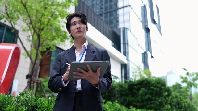 Low angle view of a young Asian businessman scrolling and writing on a digital tablet outdoors, pausing to look up in thought beside trees and a modern office building