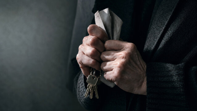 Close up of an older person holding house keys and a folded document against a dark coat