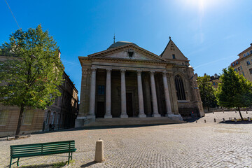 Fototapeta premium Tourists gather beneath the grand columned portico of St Pierre Cathedral on a bright sunny day in the cobblestone square of Geneva Old Town.