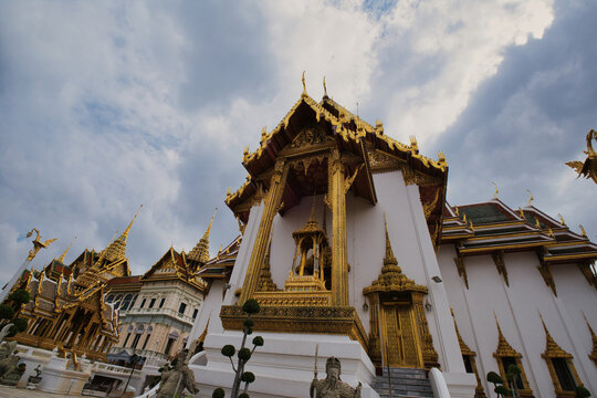 View of The Grand Palace featuring intricate gold ornamentation and traditional Thai architecture under a cloudy sky in Bangkok, Thailand.