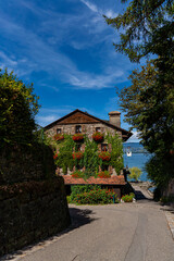 Fototapeta na wymiar A rustic stone house draped in ivy and red flowers stands at the end of a shaded lane in Yvoire, opening onto a glimpse of Lake Geneva beyond.