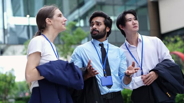Three colleagues in shirtsleeves carry their jackets and look up at the city, the bearded man pointing out a building to the businesswoman as the Asian man points upward too