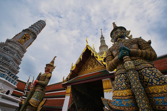 View of the ornate demon guardian statues at The Grand Palace with traditional Thai architecture and intricate golden details under a cloudy sky in Bangkok, Thailand.
