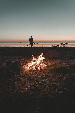 View of a person standing on the beach of Vormsi island near a glowing campfire at sunset with calm sea and rocky shoreline, Laane County, Estonia.