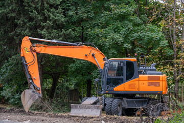 a bright orange wheeled excavator with a front dozer blade parked on a dirt construction site next to dense green trees. © Andrey