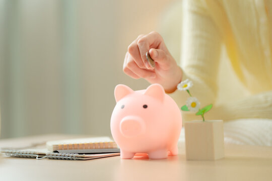 Money saving concept. Close-up of a woman's hand putting a coin into a pink piggy bank on a desk at home, with notebooks and flowers. Savings, money, coin, budgeting, finance, planning.