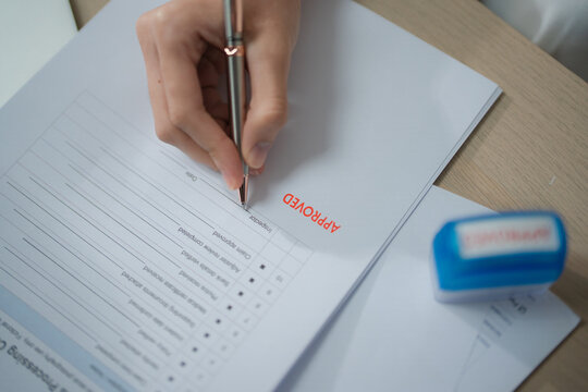 Document approval concept with an approved stamp in focus as a person signs paperwork at a desk in an office setting. Approval, document, signature, authorization, business, administration.
