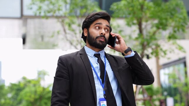 Indian businessman looks around then answers his mobile phone and begins a cheerful conversation, smiling as he talks outdoors in front of trees and a modern office building